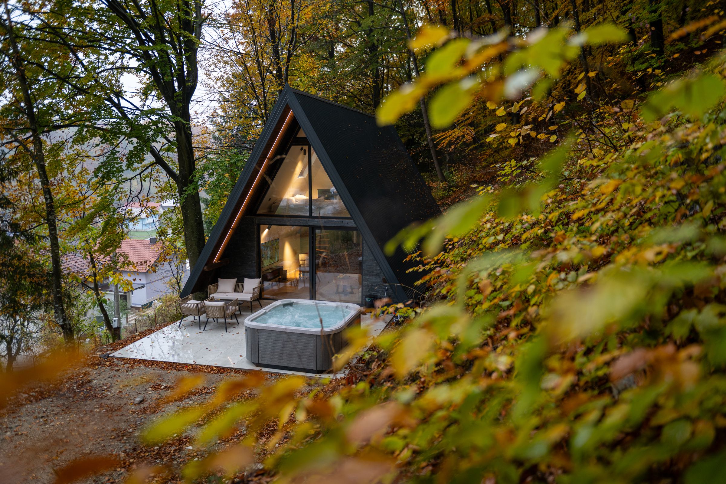 Continental Cabin seen through autumn leaves: a black A-frame cabin with a terrace and hot tub on a forest slope